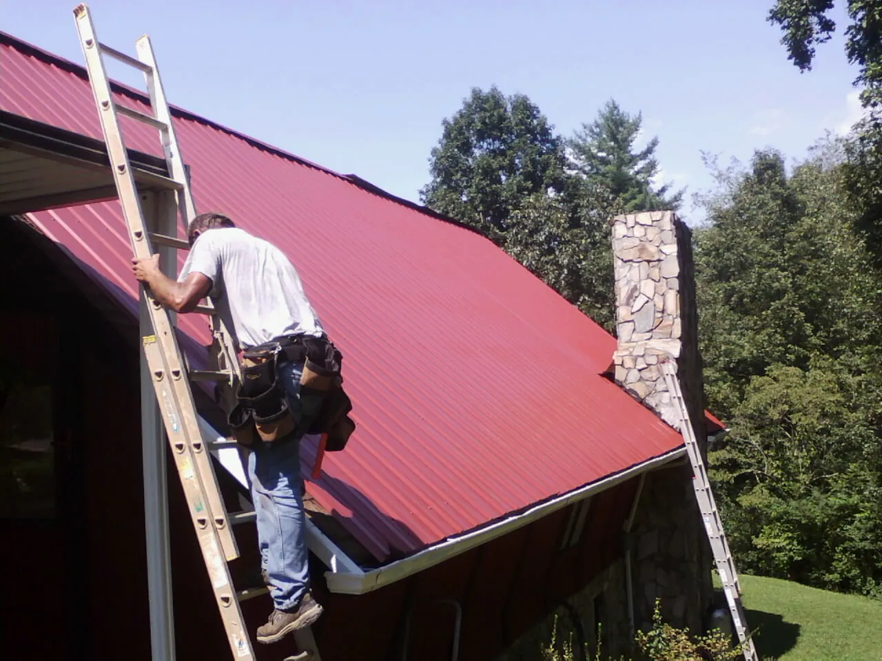 Roofer on a ladder beside a red metal roof and stone chimney — Soffit & Fascia Repair by Roofing Brothers in Black Hawk